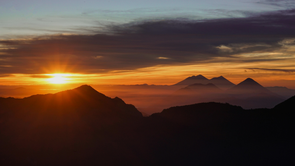 Sunrise over layered mountain ridges, with soft golden light breaking through morning clouds and illuminating a quiet landscape.